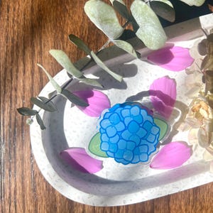 May include: A blue hydrangea flower sticker with green leaves on a white background with pink flower petals.