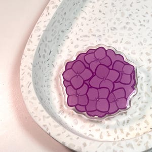 May include: A circular acrylic pin featuring a stylized illustration of a purple hydrangea flower cluster with dark purple outlines. The flower design has multiple overlapping petals and a scalloped edge, creating a distinct floral shape.
