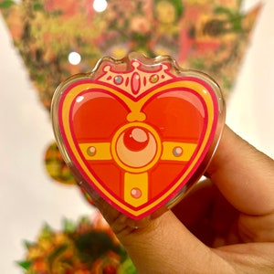 May include: A heart-shaped, clear acrylic charm with a red and yellow design. The charm features a crown, a crescent moon, and a cross-like shape. The charm is held up by a person's fingers.