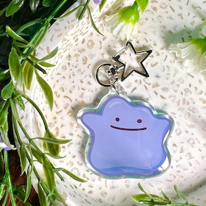 Puede incluir: Un llavero translúcido morado y azul con un personaje de dibujos animados con una cara sonriente. El llavero está unido a un amuleto plateado en forma de estrella y un anillo plateado.