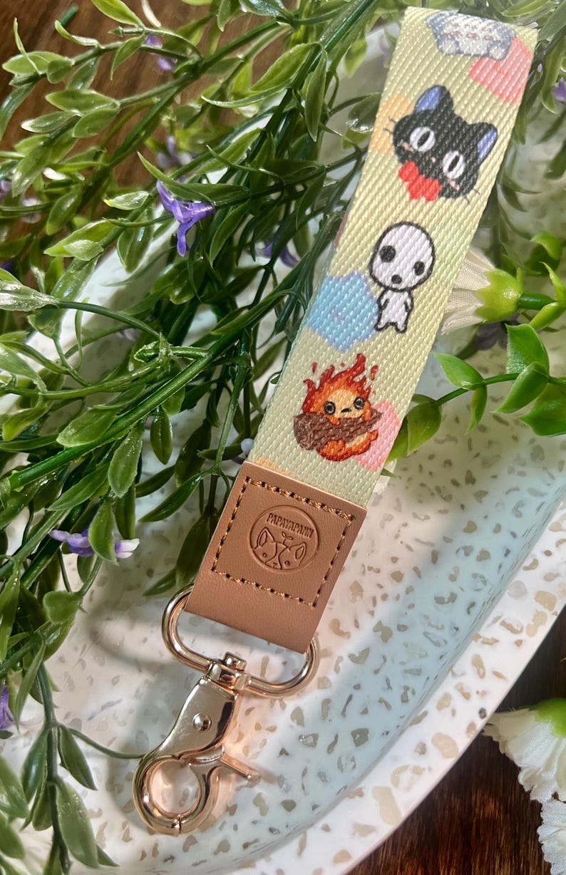 May include: A light green and white patterned key chain with a gold metal clasp. The key chain features cartoon characters from the Studio Ghibli film, My Neighbor Totoro. The key chain is attached to a brown leather strap with the word "Papillon" embossed on it.