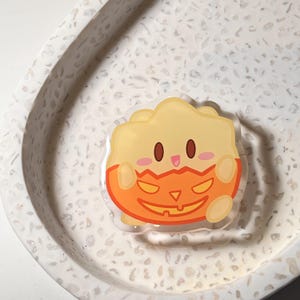 May include: A clear acrylic pin featuring a cartoon character with a yellow top and an orange jack-o'-lantern bottom. The pin has brown eyes and a smiling face. The pin is set on a white speckled tray.