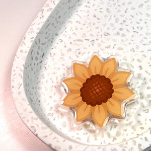 May include: A sunflower-shaped acrylic pin featuring a dark brown centre and light yellow petals. The pin has a glossy, smooth finish and is displayed on a white, teardrop-shaped tray with a speckled grey and white terrazzo pattern.