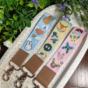 May include: Three colorful keychains with different cartoon characters. The keychains are on a white and brown speckled surface. The keychains have brown leather straps with gold metal clasps.