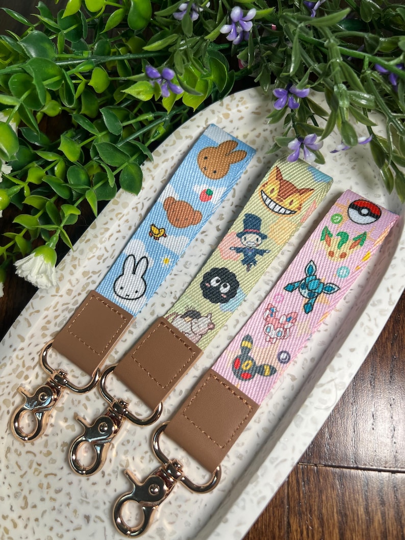 May include: Three colorful keychains with different cartoon characters. The keychains are on a white and brown speckled surface. The keychains have brown leather straps with gold metal clasps.