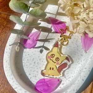 May include: A yellow and brown cartoon character keyring with a gold star charm. The keyring is on a white speckled surface.