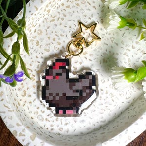May include: A grey pixelated chicken keyring with a red comb and pink legs. The keyring is attached to a gold star-shaped charm.