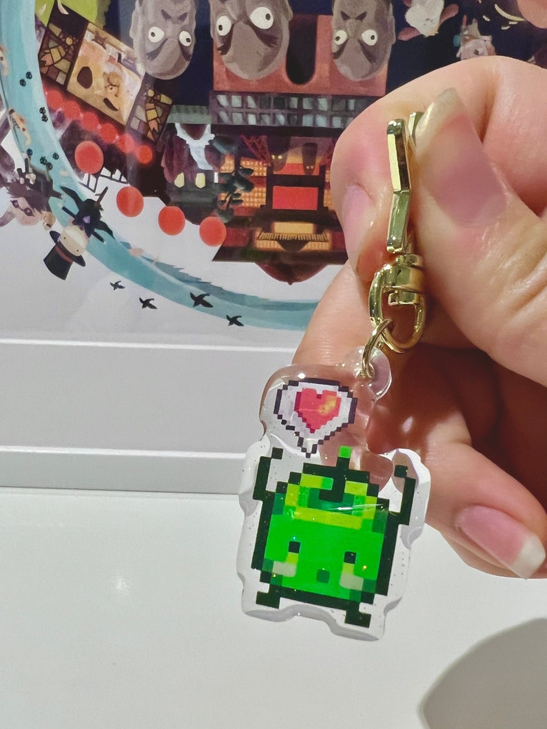 May include: A clear acrylic charm featuring a pixelated green creature with a heart-shaped thought bubble. The charm is attached to a gold-colored clasp. The background shows a colorful illustration.