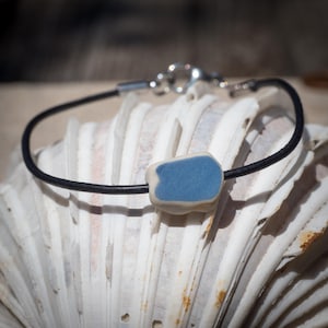 Könnte beinhalten: Ein schwarzes Lederarmband mit einer rechteckigen blauen und weißen Keramikperle. Das Armband hat einen silbernen Verschluss und wird auf einer weißen Muschelschale präsentiert. Die Perle ist hellblau.