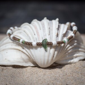 May include: A handcrafted bracelet with sea glass and beads. The bracelet features white and green beads, and a central piece of sea glass. The cord is brown, and the bracelet is displayed on a seashell.