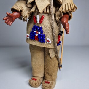 K&ouml;nnte beinhalten: Eine kleine Puppe, die ein braunes Lederoutfit mit Perlenbesatz tr&auml;gt. Die Puppe h&auml;lt einen Holzstab mit befestigten Federn.