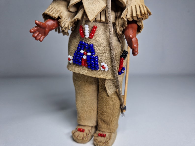 K&ouml;nnte beinhalten: Eine kleine Puppe, die ein braunes Lederoutfit mit Perlenbesatz tr&auml;gt. Die Puppe h&auml;lt einen Holzstab mit befestigten Federn.