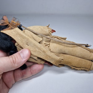 K&ouml;nnte beinhalten: Eine kleine, handgefertigte Puppe, die ein Outfit aus braunem Wildleder mit einem Federkopfschmuck tr&auml;gt. Die Puppe hat schwarze Haare und liegt auf der Seite.