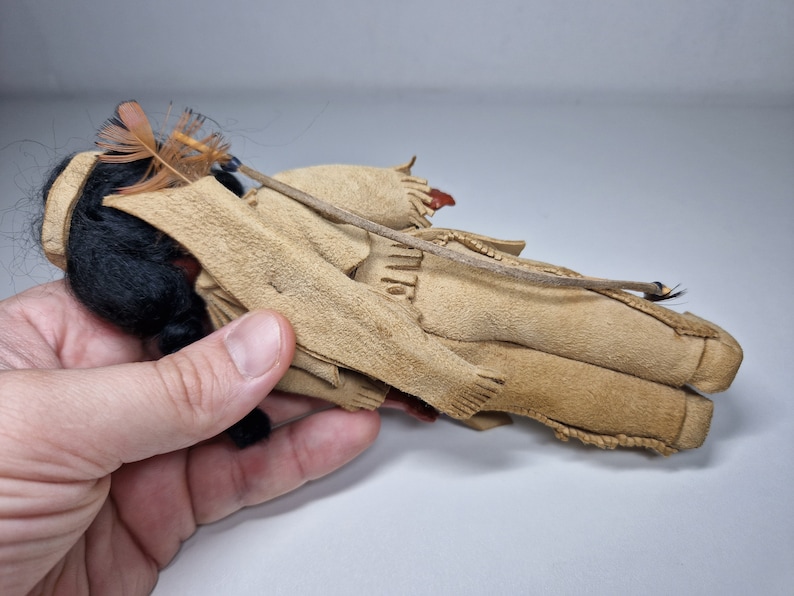K&ouml;nnte beinhalten: Eine kleine, handgefertigte Puppe, die ein Outfit aus braunem Wildleder mit einem Federkopfschmuck tr&auml;gt. Die Puppe hat schwarze Haare und liegt auf der Seite.