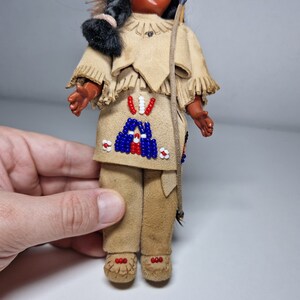K&ouml;nnte beinhalten: Eine kleine Puppe, die ein braunes Wildleder-Outfit mit Perlenbesatz tr&auml;gt. Die Puppe hat schwarze Haare und ein braunes Gesicht. Die Puppe h&auml;lt einen Bogen und Pfeil.