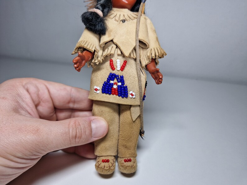 K&ouml;nnte beinhalten: Eine kleine Puppe, die ein braunes Wildleder-Outfit mit Perlenbesatz tr&auml;gt. Die Puppe hat schwarze Haare und ein braunes Gesicht. Die Puppe h&auml;lt einen Bogen und Pfeil.
