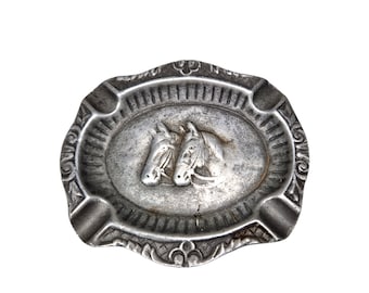 May include: Silver-toned metal ashtray with a raised oval design featuring two horse heads facing each other.