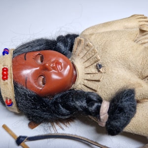 K&ouml;nnte beinhalten: Eine Indianerpuppe mit langen schwarzen Haaren, die ein braunes Wildlederkleid mit Fransen und ein Perlenband tr&auml;gt. Die Puppe hat ein bemaltes Gesicht mit geschlossenen Augen und liegt mit Bogen und Pfeil daneben auf der Seite.