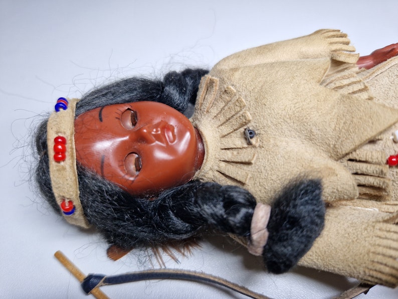 K&ouml;nnte beinhalten: Eine Indianerpuppe mit langen schwarzen Haaren, die ein braunes Wildlederkleid mit Fransen und ein Perlenband tr&auml;gt. Die Puppe hat ein bemaltes Gesicht mit geschlossenen Augen und liegt mit Bogen und Pfeil daneben auf der Seite.