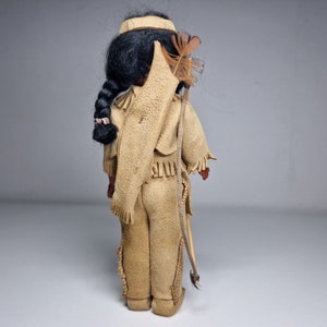 K&ouml;nnte beinhalten: Eine Vintage-Puppe eines amerikanischen Ureinwohners, die ein braunes Lederoutfit mit Fransen und einem Federkopfschmuck tr&auml;gt. Die Puppe h&auml;lt einen Bogen und Pfeil.