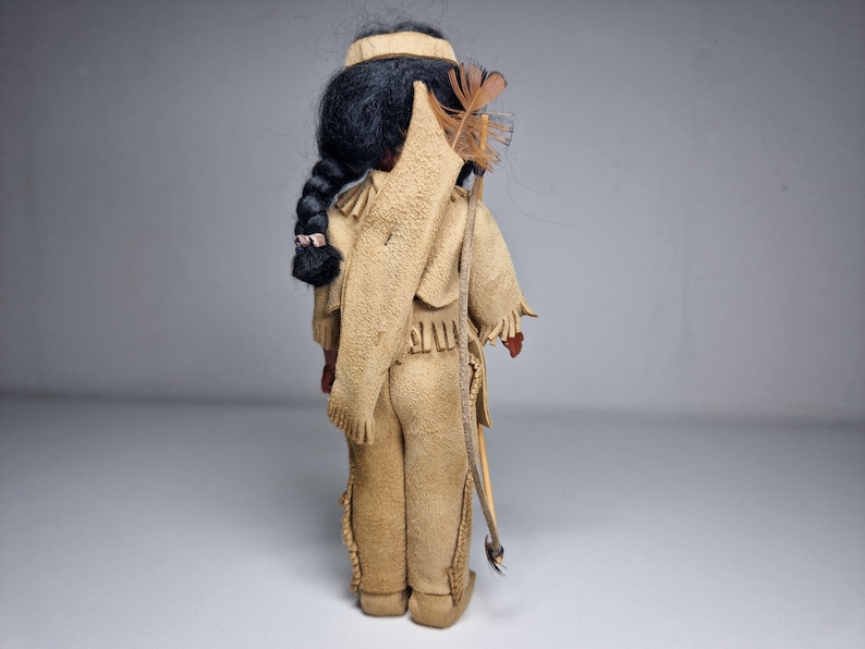 K&ouml;nnte beinhalten: Eine Vintage-Puppe eines amerikanischen Ureinwohners, die ein braunes Lederoutfit mit Fransen und einem Federkopfschmuck tr&auml;gt. Die Puppe h&auml;lt einen Bogen und Pfeil.