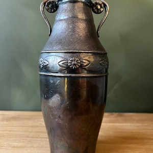 May include: A silver-toned metal vase with two curved handles and a clear glass top. The vase features a decorative band with floral designs around the middle. The vase is displayed on a wooden surface.