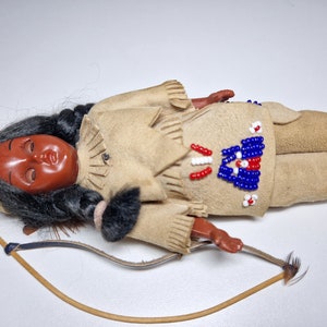 K&ouml;nnte beinhalten: Eine indianische Puppe aus Leder und Perlen. Die Puppe tr&auml;gt eine Perlenweste und hat langes schwarzes Haar. Die Puppe liegt auf dem R&uuml;cken mit Bogen und Pfeil daneben.