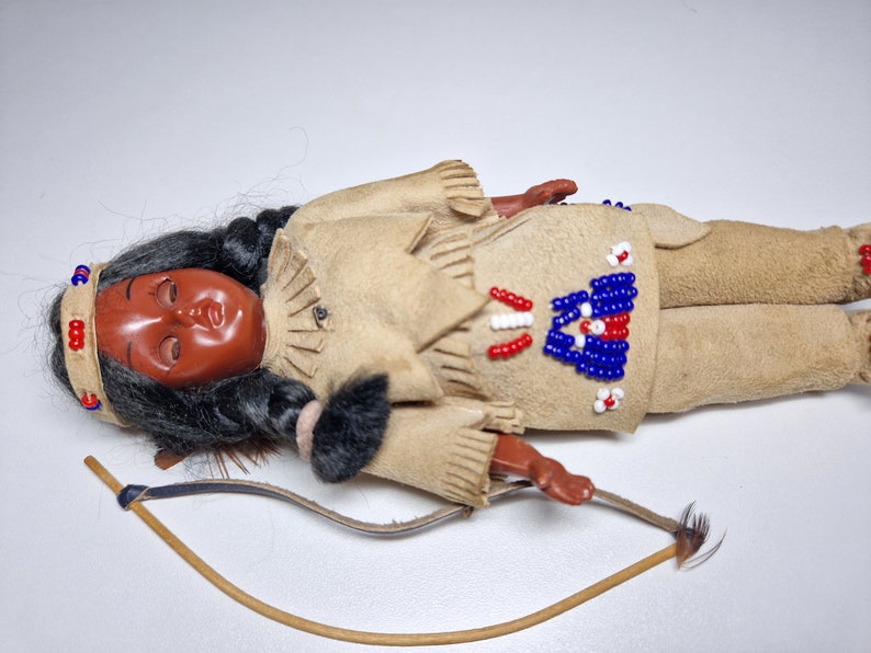 K&ouml;nnte beinhalten: Eine indianische Puppe aus Leder und Perlen. Die Puppe tr&auml;gt eine Perlenweste und hat langes schwarzes Haar. Die Puppe liegt auf dem R&uuml;cken mit Bogen und Pfeil daneben.