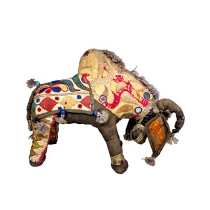 May include: A colorful, embroidered stuffed bull with a brown body and a long, curled tail. The bull is decorated with intricate patterns in shades of red, yellow, green, and blue.