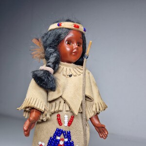 K&ouml;nnte beinhalten: Eine Vintage-Puppe eines amerikanischen Ureinwohners mit schwarzen Haaren, braunen Augen und einem Perlenkopfschmuck. Die Puppe tr&auml;gt ein Fransenkleid aus Wildleder mit Perlenverzierungen.