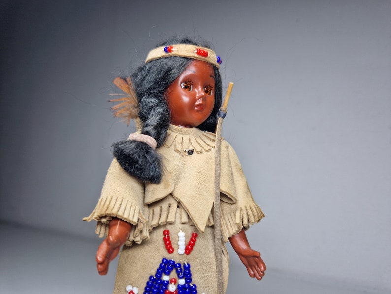 K&ouml;nnte beinhalten: Eine Vintage-Puppe eines amerikanischen Ureinwohners mit schwarzen Haaren, braunen Augen und einem Perlenkopfschmuck. Die Puppe tr&auml;gt ein Fransenkleid aus Wildleder mit Perlenverzierungen.