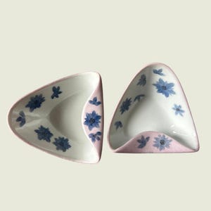 Puede incluir: Dos platos triangulares de cerámica. Los platos tienen un interior blanco y un borde exterior rosa claro. Los patrones florales azules decoran el interior y el exterior. Los platos son probablemente para servir aperitivos o porciones pequeñas.
