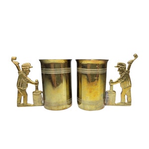 May include: Two gold-tone metal mugs featuring a design of a person holding a paint roller. The mugs are set against a white background.