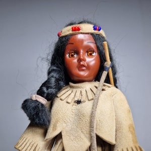 K&ouml;nnte beinhalten: Eine Vintage-Puppe eines amerikanischen Ureinwohners mit schwarzen Haaren, brauner Haut und orangefarbenen Augen. Die Puppe tr&auml;gt ein braunes Wildleder-Outfit mit Fransen und einem Perlenband.