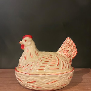 Könnte beinhalten: Ein cremefarbener Keramikbehälter in Form eines Huhns mit roten Akzenten. Das Huhn sitzt auf einem nestartigen Sockel, mit rotem Kamm und Kehllappen. Das Design weist detaillierte Feder- und Neststrukturen auf.