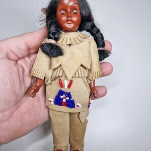 K&ouml;nnte beinhalten: Eine kleine, antike Puppe, die in einem traditionellen indianischen Kost&uuml;m gekleidet ist. Die Puppe hat schwarze Haare in Z&ouml;pfen, ein braunes Gesicht und tr&auml;gt ein braunes Wildleder-Outfit mit Perlenbesatz.