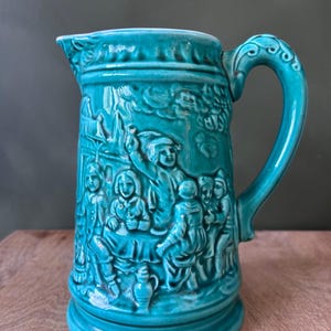 Puede incluir: Una jarra de cerámica color verde azulado con un diseño en relieve detallado de un grupo de personas reunidas en una taberna. El recipiente tiene un asa curva, un pico ancho y un acabado brillante que resalta las figuras moldeadas.