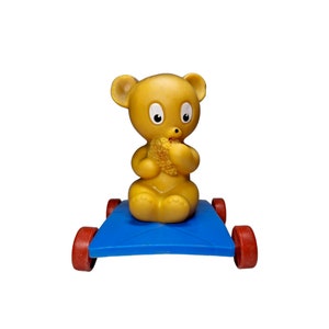 Puede incluir: Un oso de peluche amarillo con un carro azul. El oso sostiene un panal de miel de juguete amarillo. El carro tiene ruedas rojas.