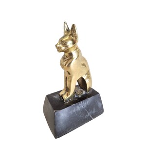 May include: A gold-colored metal cat figurine sitting upright on a dark gray rectangular base. The cat has pointed ears and is facing to the left. The base appears to be made of stone.