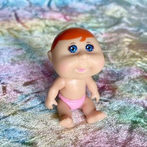 Könnte beinhalten: Eine kleine Baby-Puppe aus Kunststoff mit heller Haut, rotem Haar und leuchtend blauen Augen. Die Puppe trägt eine rosa Windel. Die Puppe sitzt auf einer mehrfarbigen, strukturierten Oberfläche.