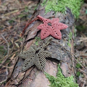 Peut inclure: Deux pendentifs en macramé en forme de feuille, l'un de couleur vert foncé et l'autre de couleur rose clair, sont présentés sur un tronc d'arbre recouvert de mousse.
