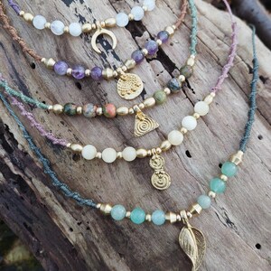 Peut inclure: Cinq colliers superposés avec des perles de différentes couleurs et des breloques dorées. Les colliers présentent une lune croissante, un motif circulaire, un triangle avec une spirale, deux cercles et une feuille.