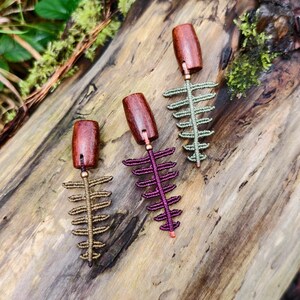 May include: Three handmade macrame earrings with wooden tops and fern-like designs in shades of brown, purple, and green. The earrings are displayed on a weathered wooden surface.