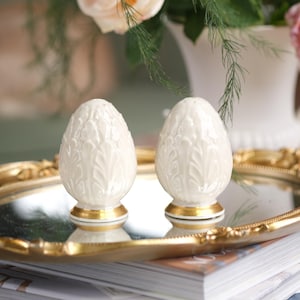 Puede incluir: Dos saleros y pimenteros blancos en forma de huevo, con un diseño de hojas y ribete dorado, exhibidos en una bandeja con espejo y borde dorado. La bandeja está sobre una pila de libros, con un arreglo floral borroso en el fondo.