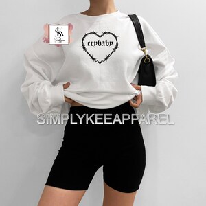 May include: White sweatshirt with a black heart-shaped design featuring barbed wire and the word "crybaby." The sweatshirt is paired with black biker shorts. A black purse hangs from the shoulder. The brand name "SIMPLYKEEAPPAREL" is displayed.
