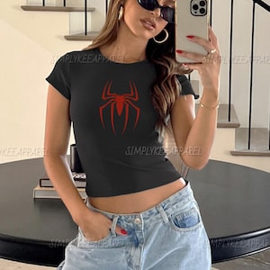 May include: Black crop top featuring a red spider design. The top has a round neck and short sleeves. The text "SIMPLYKEE APPAREL" is printed on the shirt. The model is wearing blue jeans and sunglasses.