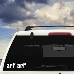 以下が含まれることがあります： 大胆で様式化されたフォントで「arf arf」というテキストが書かれた白い車の窓用ステッカー。白い車のリアウィンドウに貼られており、背景には曇り空が広がっています。Simply Kee Apparelのステッカーです。