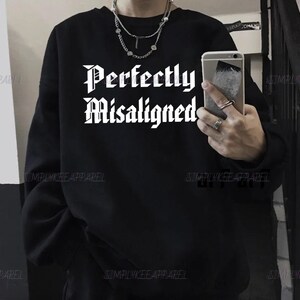 May include: Black sweatshirt with the text "Perfectly Misaligned" in a gothic font. The sweatshirt has a crew neck and long sleeves. The brand name "SIMPLYKEE APPAREL" is printed at the bottom of the sweatshirt.