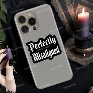 Pode incluir: Capa de telefone cinza com a frase "Perfectly Misaligned" em fonte gótica. A capa tem um acabamento brilhante e um adesivo em preto e branco. A marca "SIMPLYKEE APPAREL" é impressa ao redor da capa. Uma vela roxa acesa ao fundo.