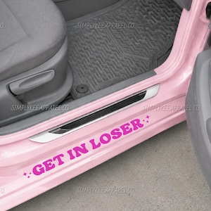 May include: A bright pink car door sill decal featuring the bold, magenta text "GET IN LOSER" flanked by two small four-pointed star icons. The sticker is applied to the lower interior frame of a light pink vehicle, positioned below the gray seat.